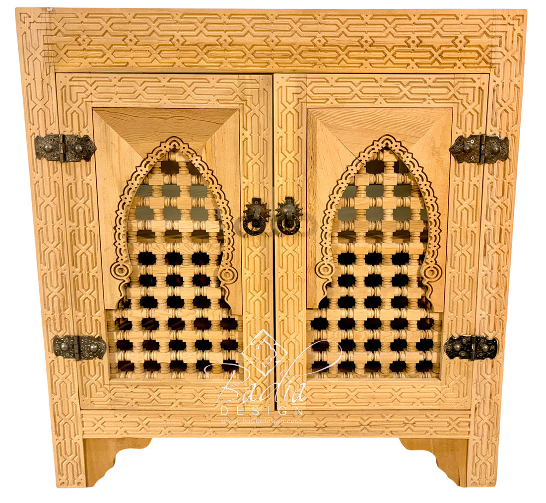 An ornate light wood cabinet with two doors, featuring intricate geometric carvings, arched lattice cutouts on the doors, and dark metal hinges and pulls, shown against a plain white background.