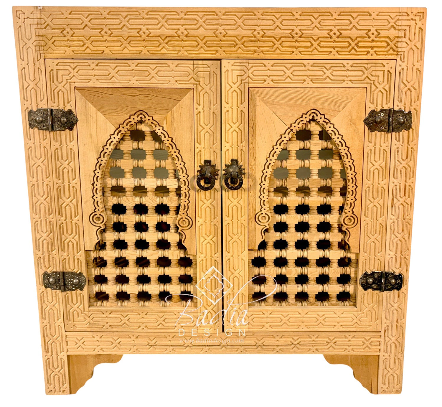An ornate light wood cabinet with two doors, featuring intricate geometric carvings, arched lattice cutouts on the doors, and dark metal hinges and pulls, shown against a plain white background.