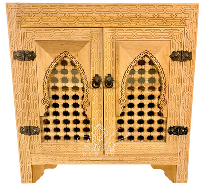 An ornate light wood cabinet with two doors, featuring intricate geometric carvings, arched lattice cutouts on the doors, and dark metal hinges and pulls, shown against a plain white background.