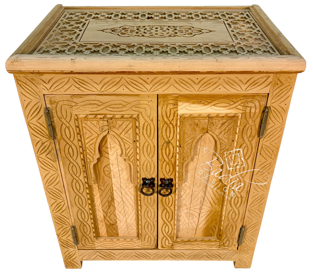 A light natural wood cabinet with intricate hand-carved geometric and floral patterns, featuring two doors with dark metal ring pulls and hinges, set against a plain white background.