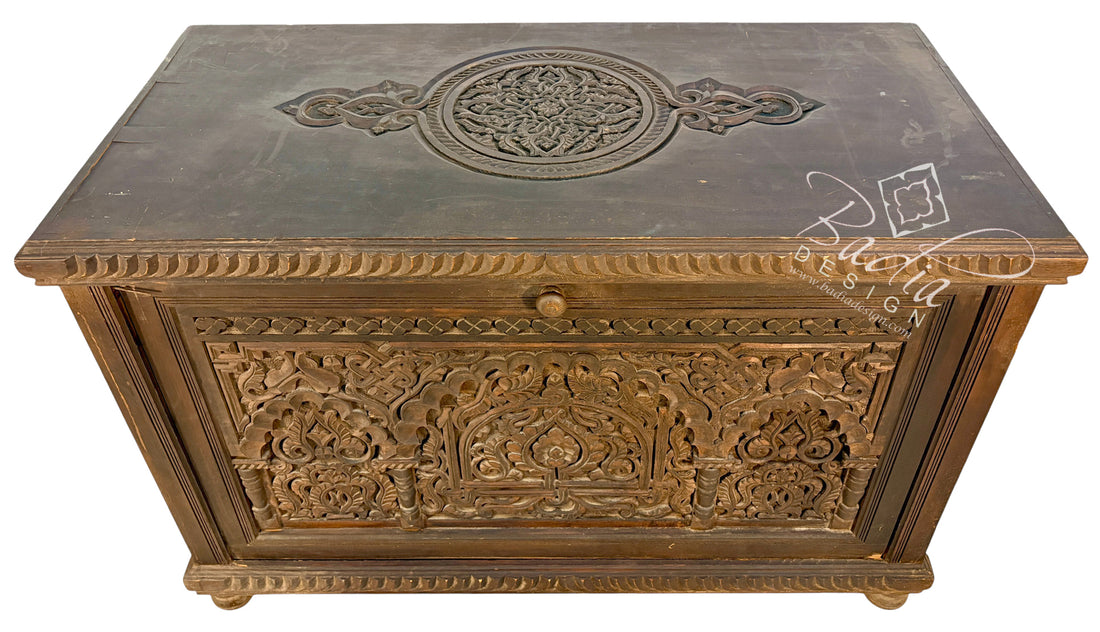 An ornate carved wooden chest or trunk with intricate floral and geometric patterns, featuring a single round knob and a dark top surface with a circular relief design.