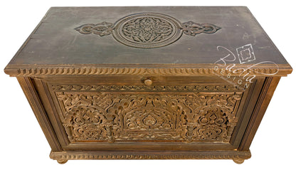 An ornate carved wooden chest or trunk with intricate floral and geometric patterns, featuring a single round knob and a dark top surface with a circular relief design.