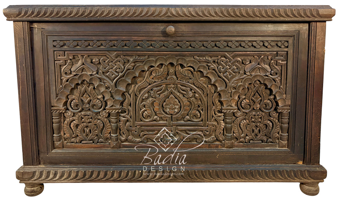 An ornate dark brown carved wooden chest or cabinet with intricate floral and arch motifs, shown against a plain white background. A &