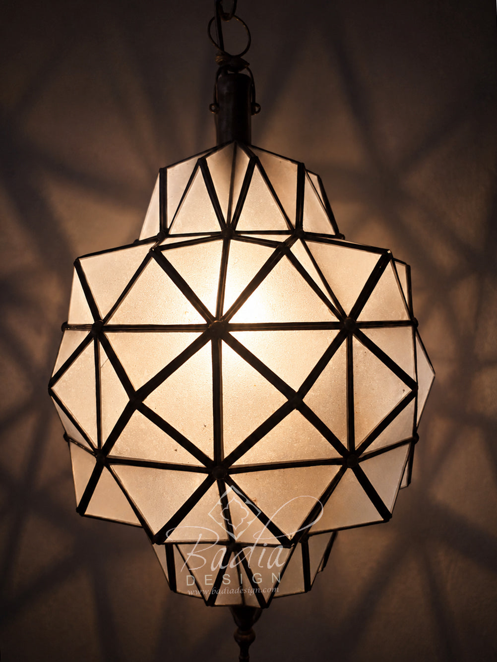 A faceted geometric pendant light with a warm glowing frosted glass shade and dark metal frame, hanging against a dark wall with soft shadow patterns cast around it.