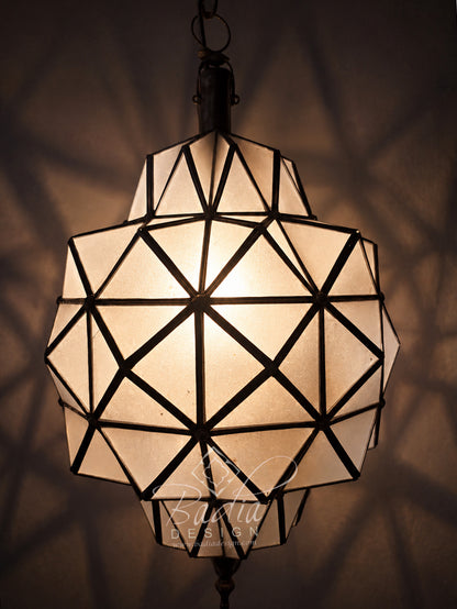 A faceted geometric pendant light with a warm glowing frosted glass shade and dark metal frame, hanging against a dark wall with soft shadow patterns cast around it.