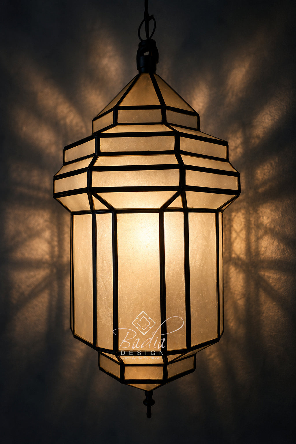 A hanging Moroccan-style lantern with geometric glass panels and a warm glowing light inside, casting dramatic shadow patterns on a dark background.