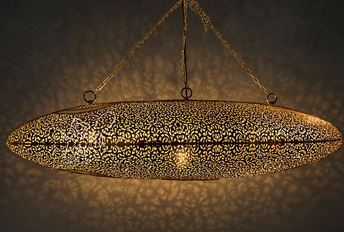A large oval Moroccan-style hanging pendant lamp with an intricate laser-cut metal pattern, casting warm golden light and shadow patterns on a textured wall.